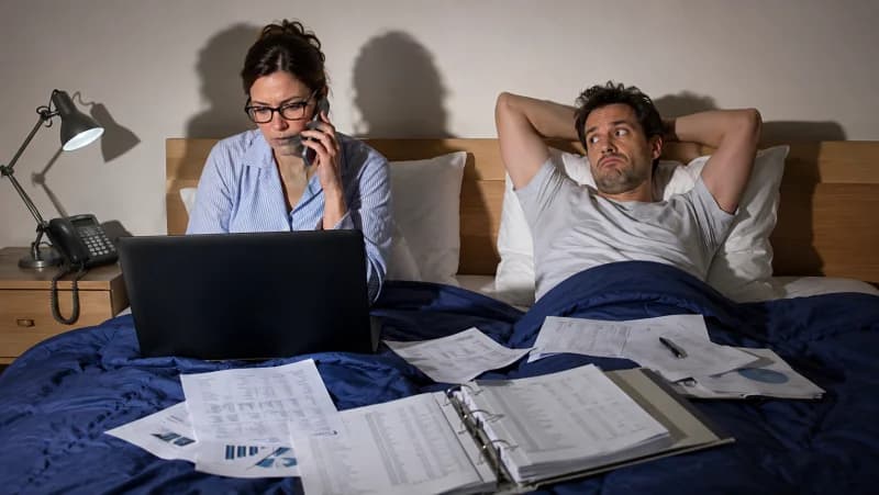 A couple lies in bed at night as the woman works on her laptop and takes a phone call, surrounded by paperwork, while her frustrated partner looks on.